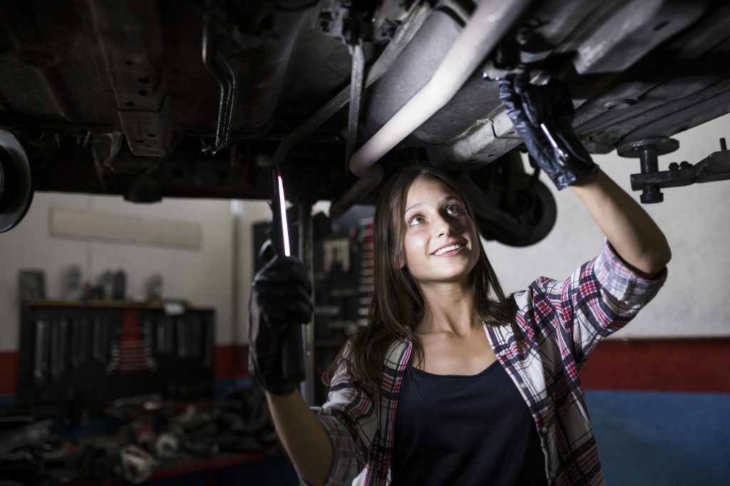 Smiling woman with lamp fixing car standing under bottom in car repair service